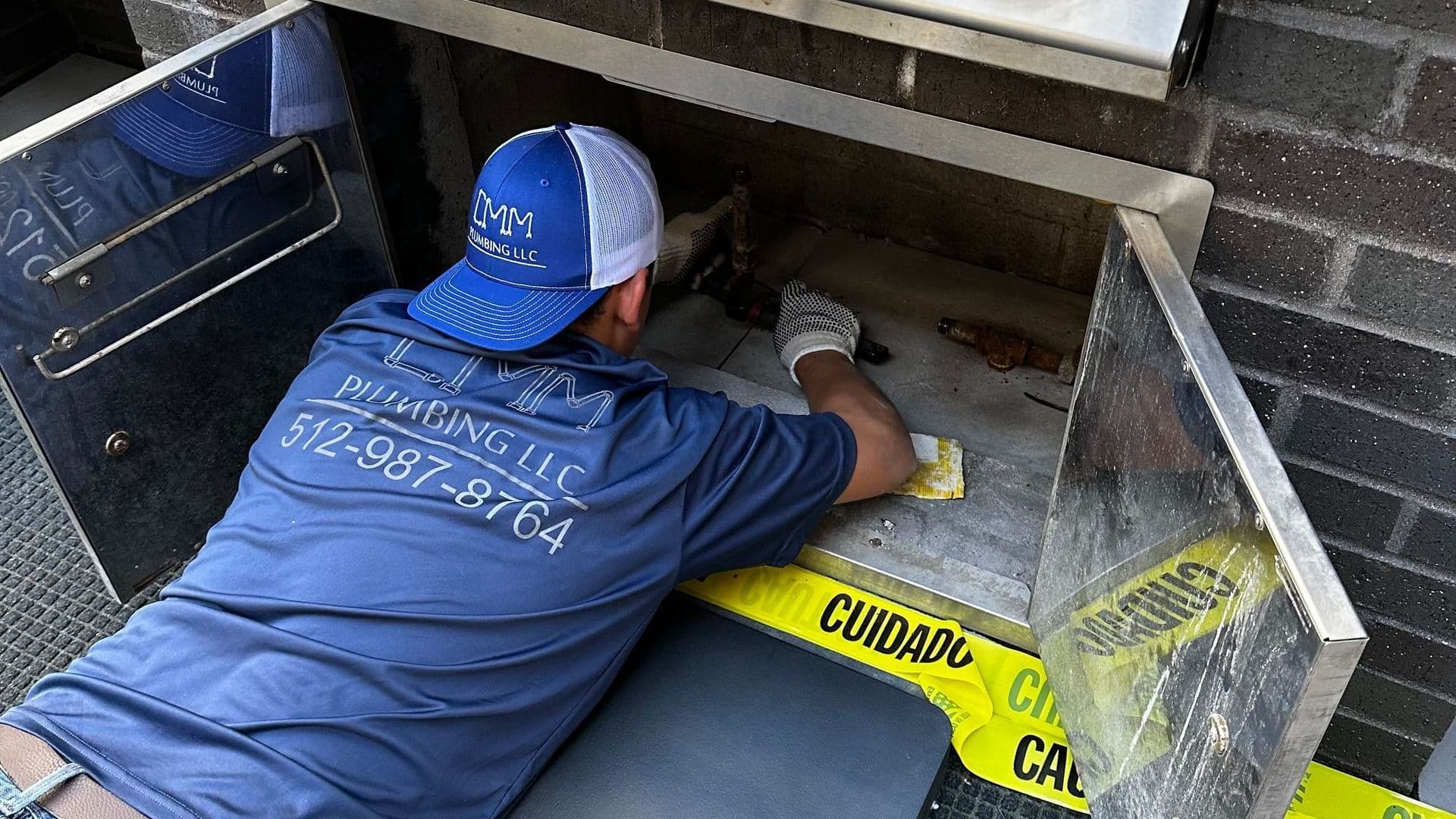 A person wearing a blue shirt and hat cleans a built-in outdoor grill with yellow caution tape nearby.