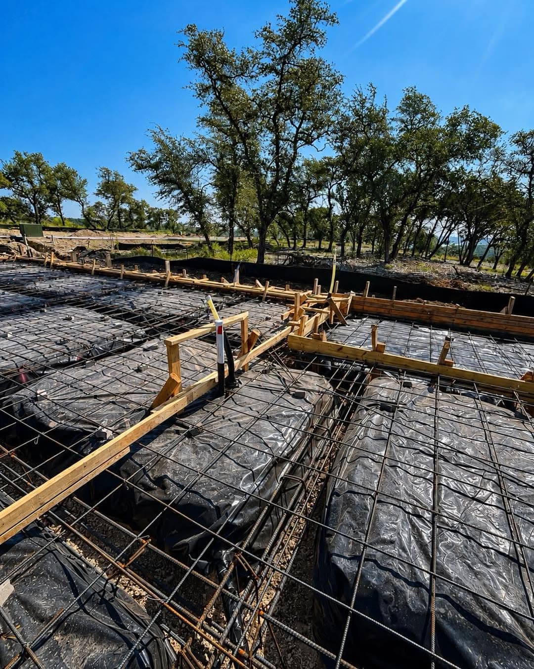 Rebar tied over a vapor barrier with plumbing lines run beneath it, framed by wood forms on a Central Texas home site.