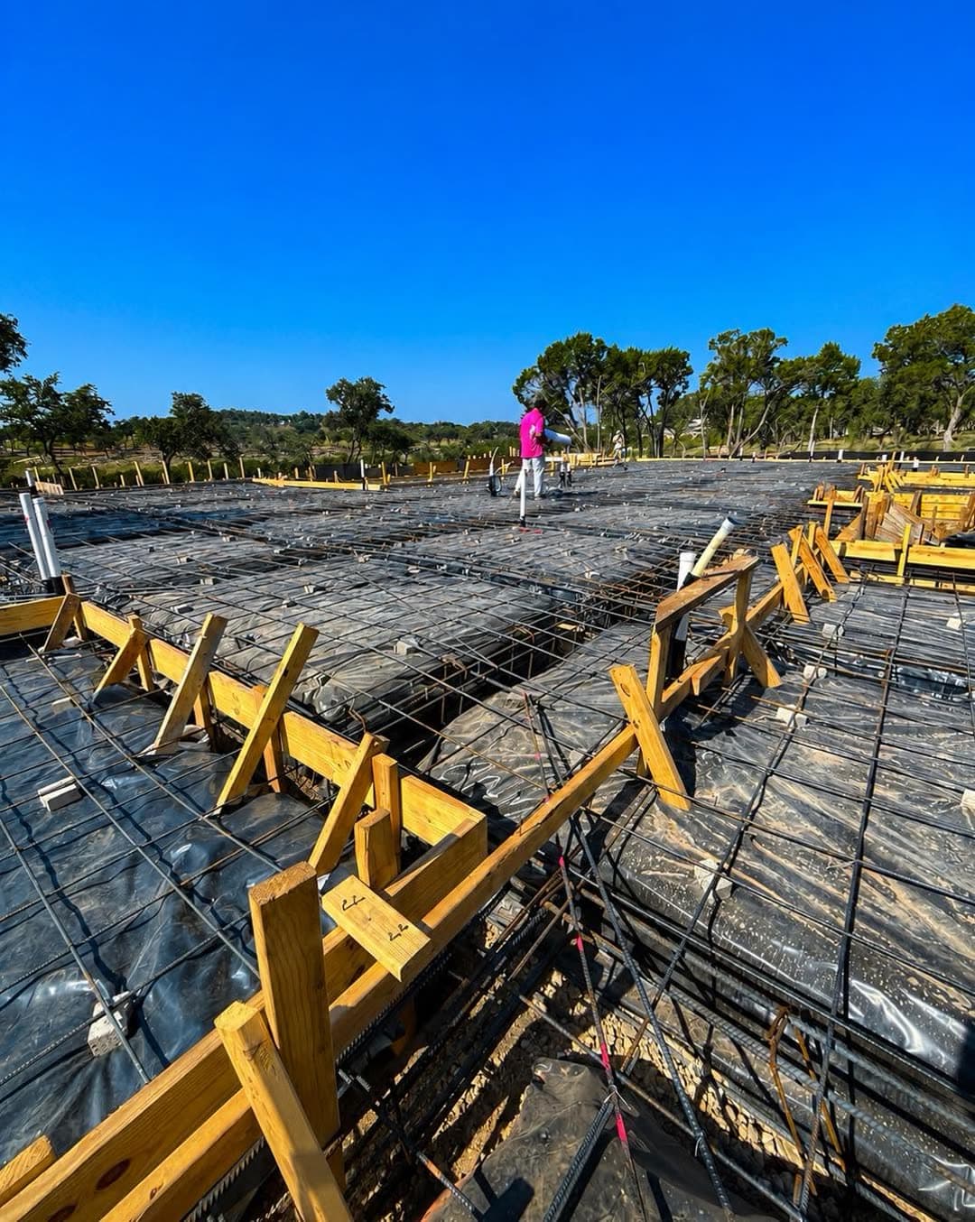 Wood bracing and rebar across a prepared slab with under-slab plumbing complete, a crew member working in the distance.